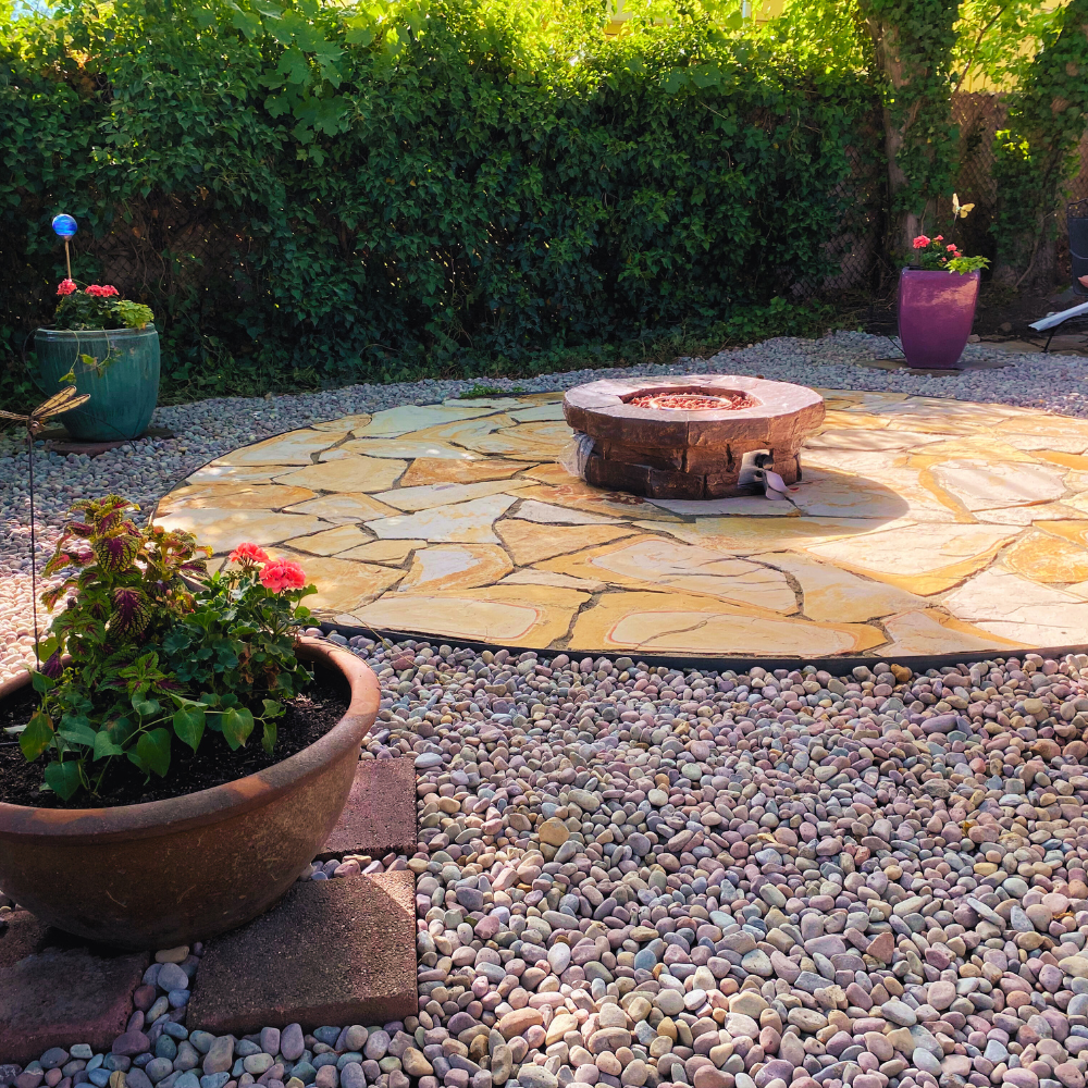 Flagstone patio in a round shape, in a landscaping scene, with custom fire pit, planter pots with flowers. Rainbow pebbles, ivy