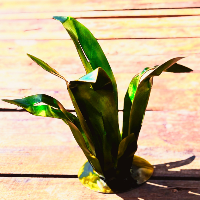 Green and yellow painted metal in the shape of leaves, welded to a round yellow base, forms an agave shaped plant. Fake plant, metal art, for garden. Displayed on a wooden pathway with a shadow to the right.