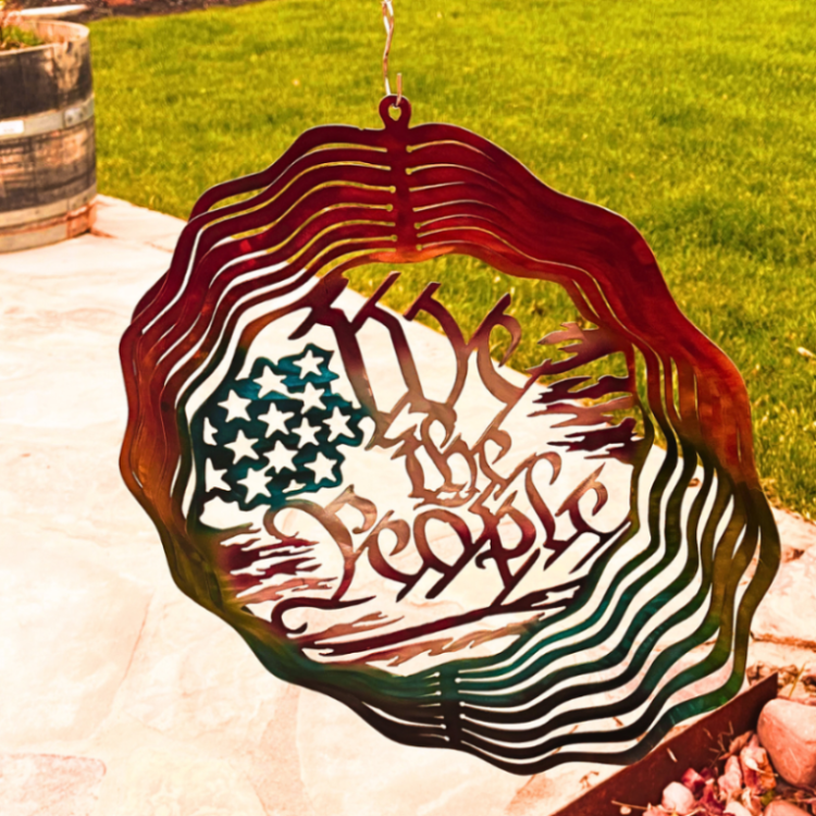 A metal wind spinner with a hoo at the top. 7 rings extend and catch wind that make it spin on the 360 degree hook. Center says "we the people" and has tattered flag shapes and stars that represent the USA flag. Grass and patio in the background